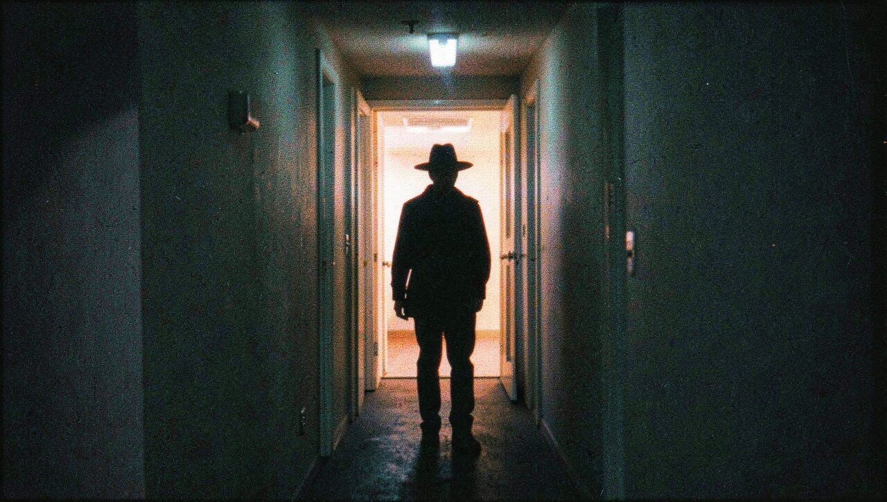 A featureless silhouette at the end of an apartment hallway, lit by sodium vapor from an open door. Head, shoulders, torso. Nothing else.