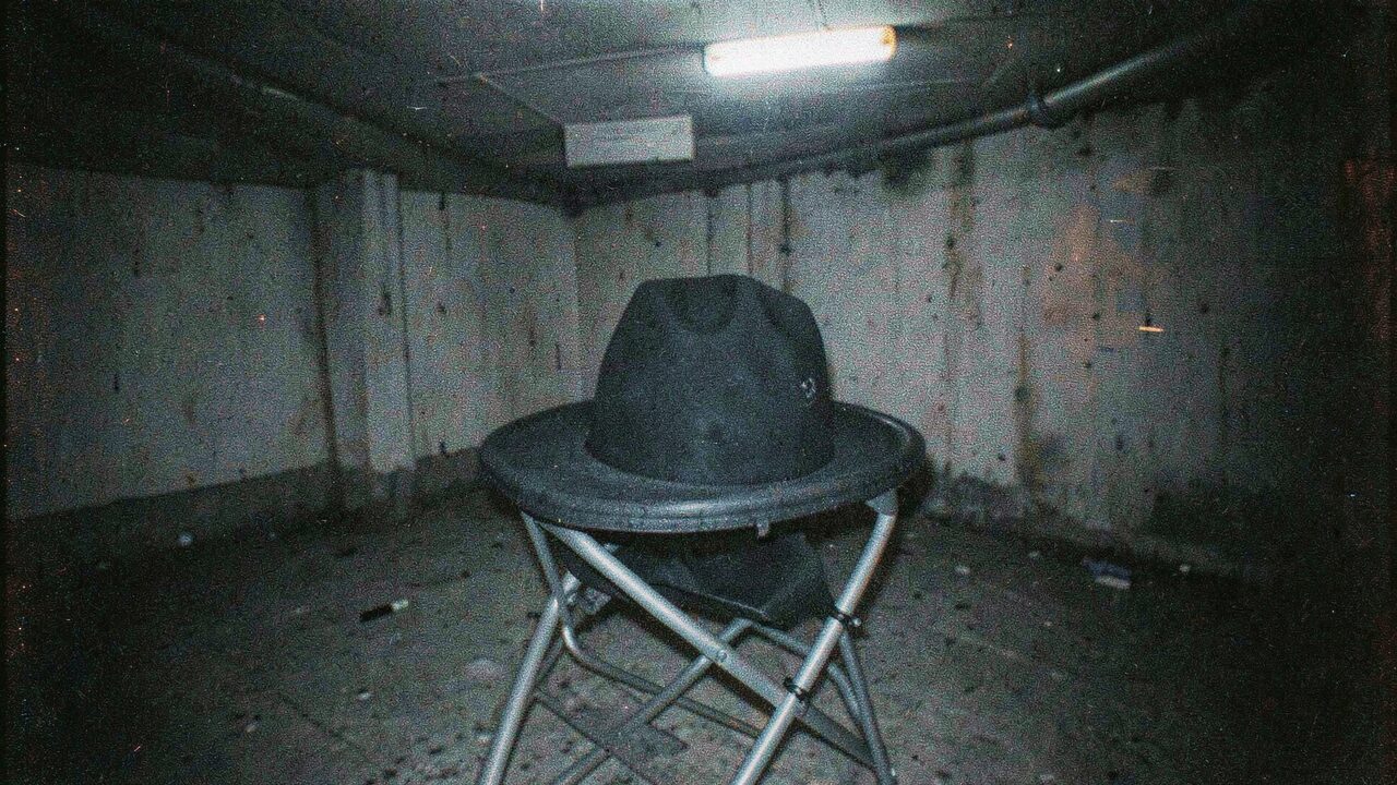 A black wide-brimmed hat on a folding chair in a dim basement, caught in flash. The accessory, without its owner.