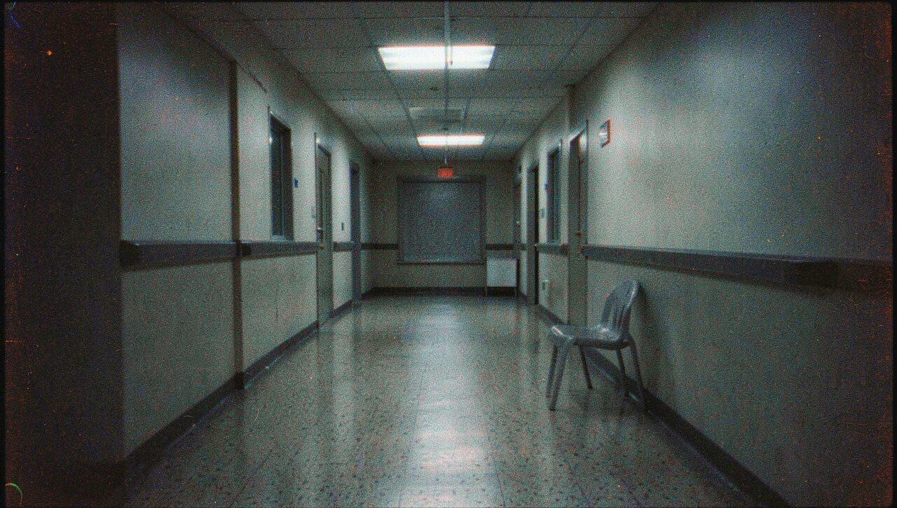 An empty hospital corridor at night, fluorescent light on linoleum, a single plastic chair against the wall. The kind of place he gets reported, eventually.