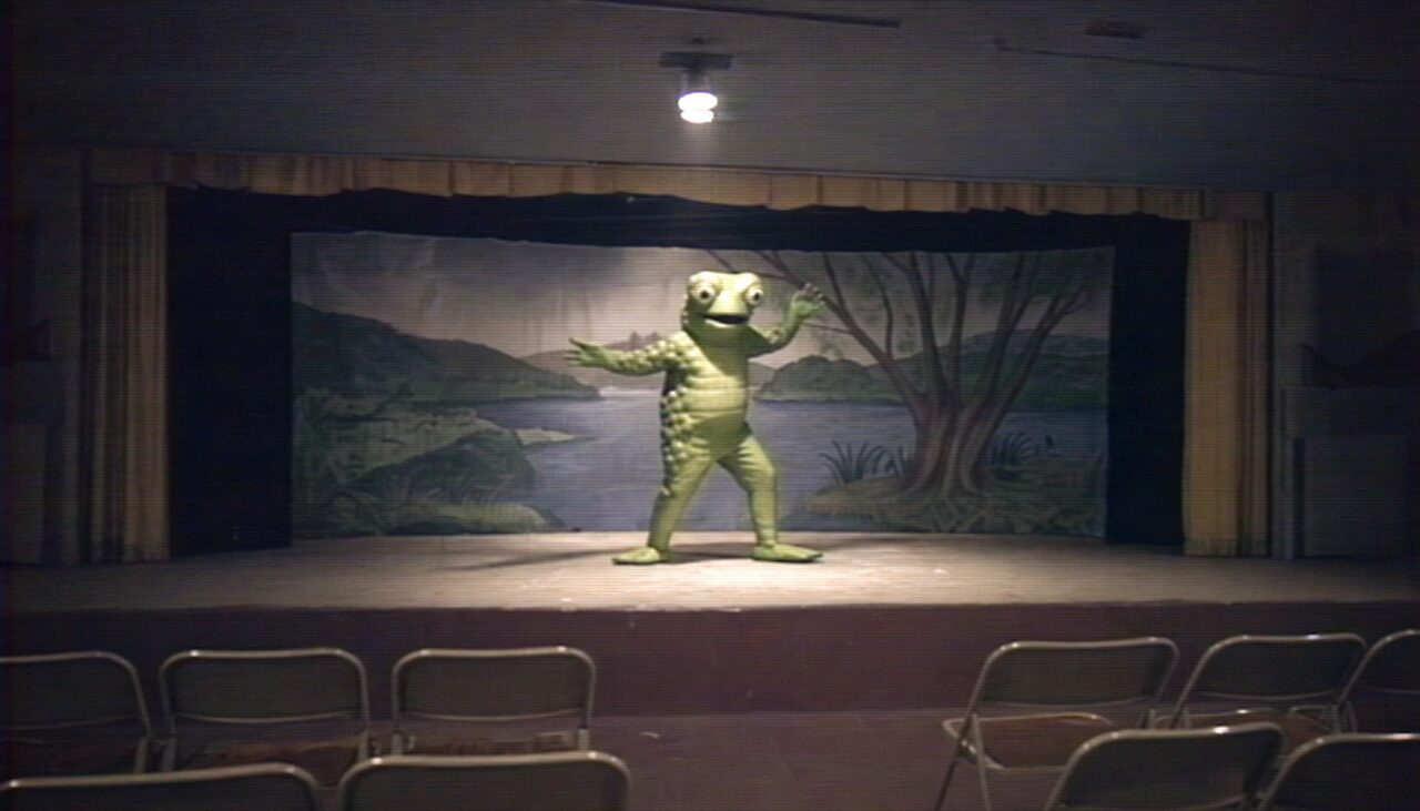 A performer in a handmade green frog costume, frozen mid-pose under a single spotlight, a hand-painted riverbank behind them.