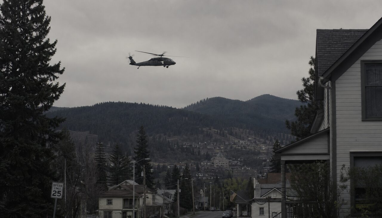 A low military helicopter against flat grey overcast over forested hills, seen from a residential street.