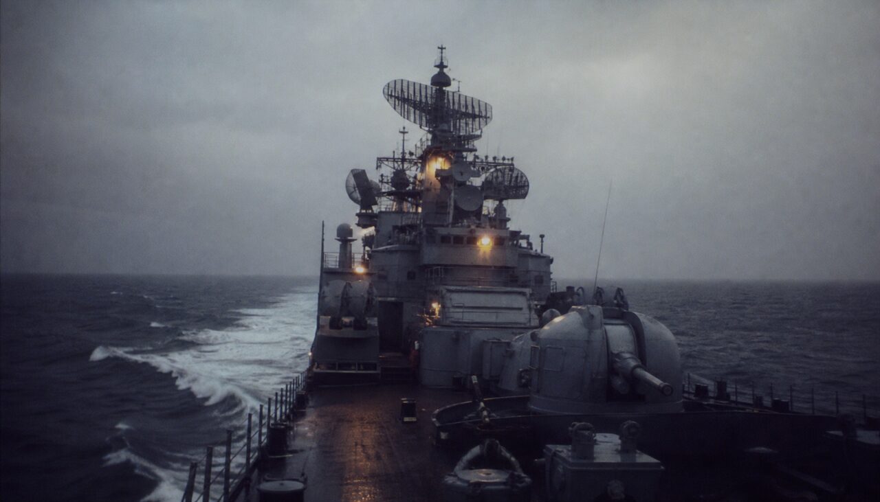 A guided-missile cruiser cutting through dark Pacific swells, radar arrays turning against a flat overcast. No land in any direction.