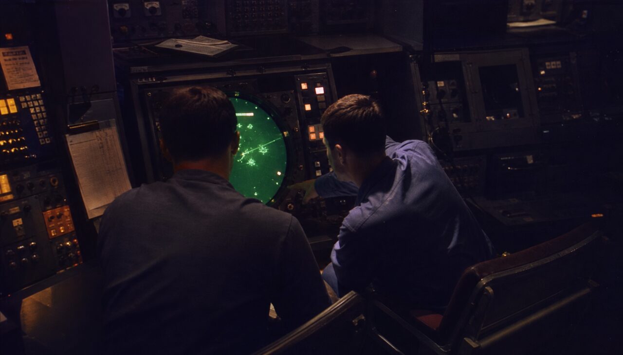 Two radar operators leaning toward the same scope in a darkened Combat Information Center, faces lit green from below.