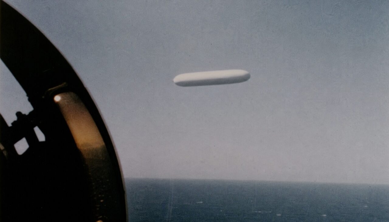 A smooth white oblong hanging motionless above the flat Pacific, seen through the canopy frame. No wings, no markings, no seams.